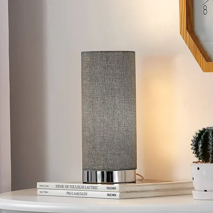 Lindby Tischleuchte Ronja - alu / grau / zink, E14 - Graue Stoff-Tischlampe mit Chromfuß auf einem Tisch, neben Büchern.