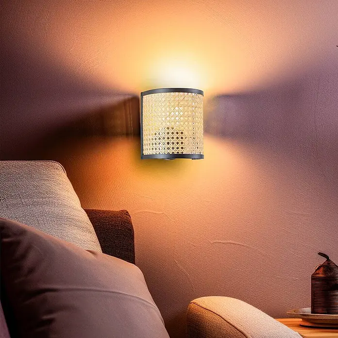 Lindby Wandleuchte Yaelle - Holz hell, E27 - Wandleuchte mit schwarzem Rahmen und Rattan-Geflecht, beleuchtet eine warm getönte Wand über einem Sofa.