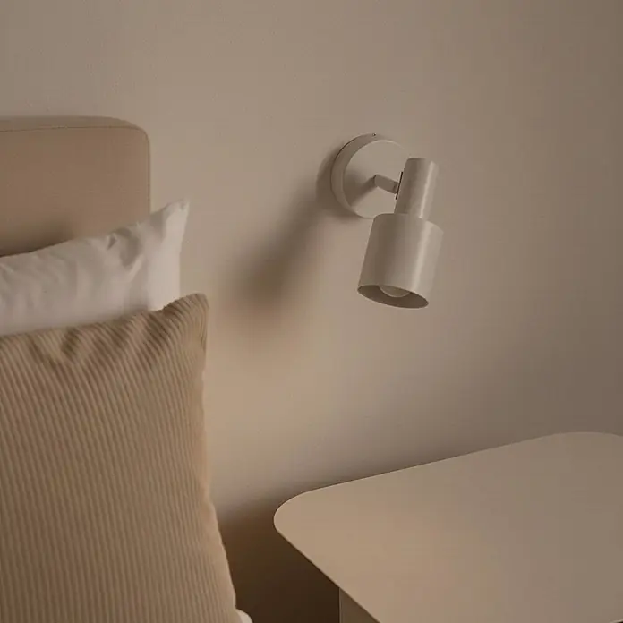 Lindby Wandstrahler Ovelia - creme / amber, creme / amber, E27 - Ein hellbeiger Wandstrahler ist über einem passenden Nachttisch neben einem Bett positioniert.