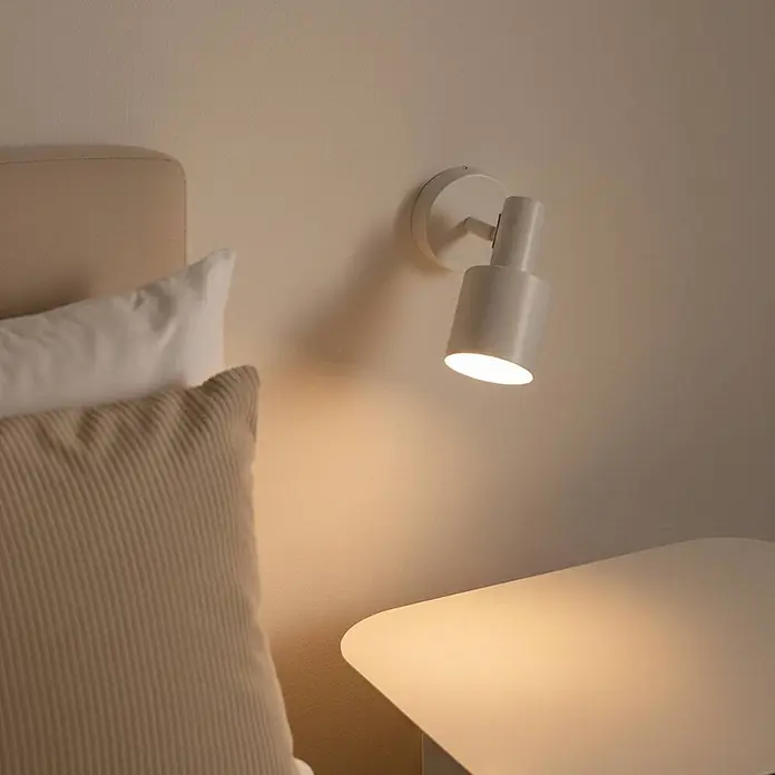 Lindby Wandstrahler Ovelia - creme / amber, creme / amber, E27 - Weißer Wandstrahler neben einem Bett, der einen hellen Nachttisch beleuchtet und ein warmes Licht erzeugt.