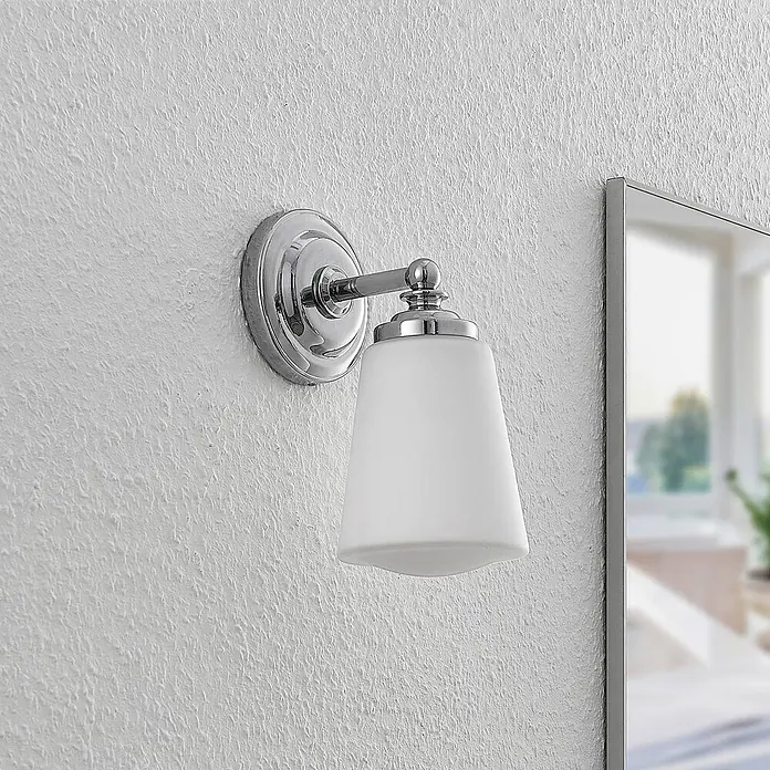 Arcchio Wandleuchte Mahdi - chrom / silber, E14 - Chrom-Wandleuchte mit mattweißem Glasschirm, an einer strukturierten Wand neben einem Spiegel montiert.