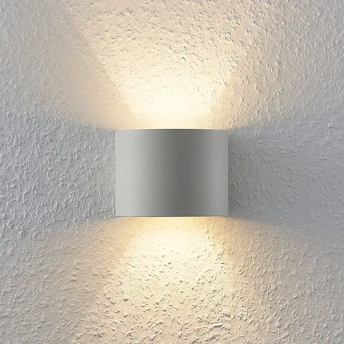 Arcchio Wandleuchte Zuzana - L x B x H: 11,2 x 13 x 9,7 cm, weiß / opal, G9 - Eine moderne graue zylindrische Wandleuchte wirft warmes Licht nach oben und unten auf eine strukturierte weiße Wand.