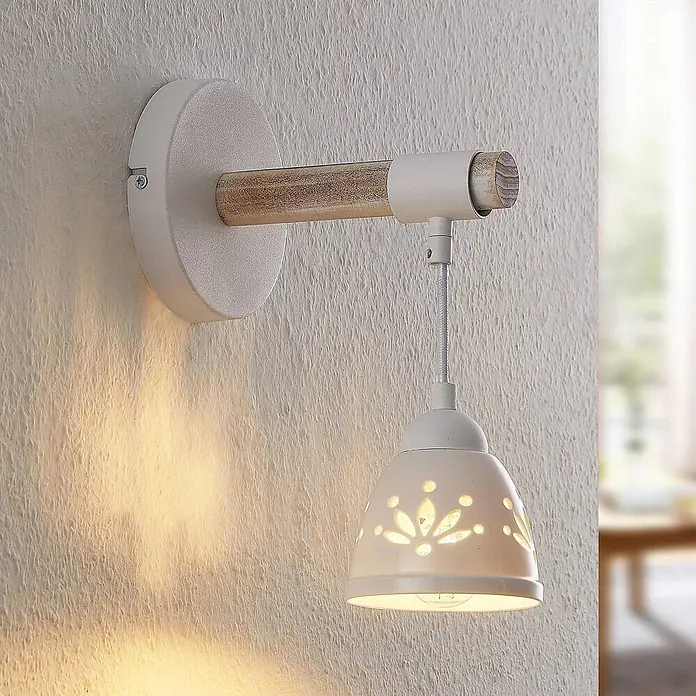 Lindby Wandleuchte Pimana - Holz hell, weiß / opal, E14 - Weiße Wandleuchte mit Holzarm und Keramikschirm mit Blumenmuster-Ausschnitten, die ein warmes Licht wirft.