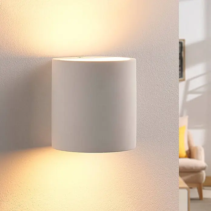 Lindby Wandleuchte Krista - L x B x H: 10 x 10,5 x 10 cm, weiß / opal, G9 - Weiße zylindrische Wandleuchte an einer strukturierten Wand, die warmes Licht nach oben und unten abgibt.