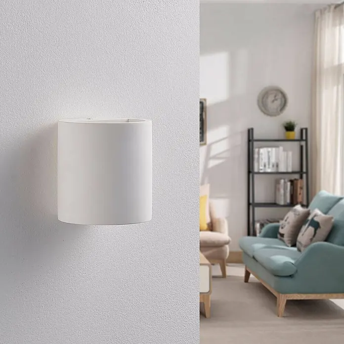 Lindby Wandleuchte Krista - L x B x H: 10 x 10,5 x 10 cm, weiß / opal, G9 - Weiße zylindrische Wandleuchte, an einer strukturierten Wand montiert, teilweise einen unscharfen Wohnbereich verdeckend.