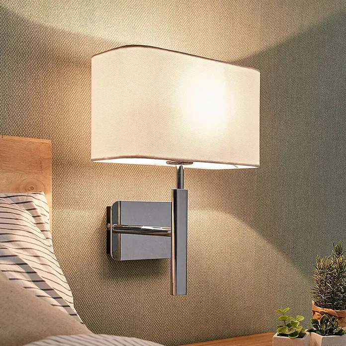 Lucande Wandleuchte Jettka - chrom / silber, E27 - Eine Wandleuchte mit weißem, rechteckigem Stoffschirm und Chromfuß beleuchtet eine texturierte Wand neben einem Bett.