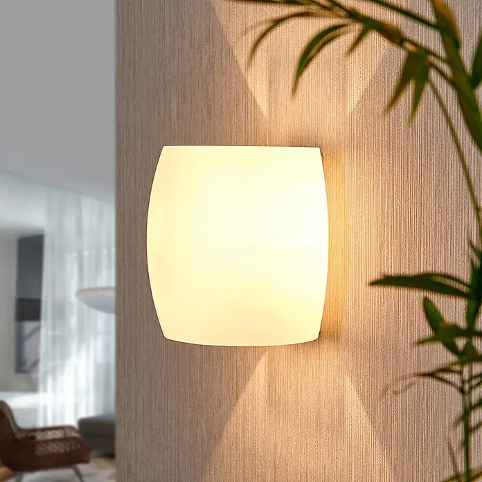 Lucande Wandleuchte Lusine - weiß / opal, G9 - Gebogene weiße Wandleuchte wirft warmes, diffuses Licht auf eine strukturierte Wand.