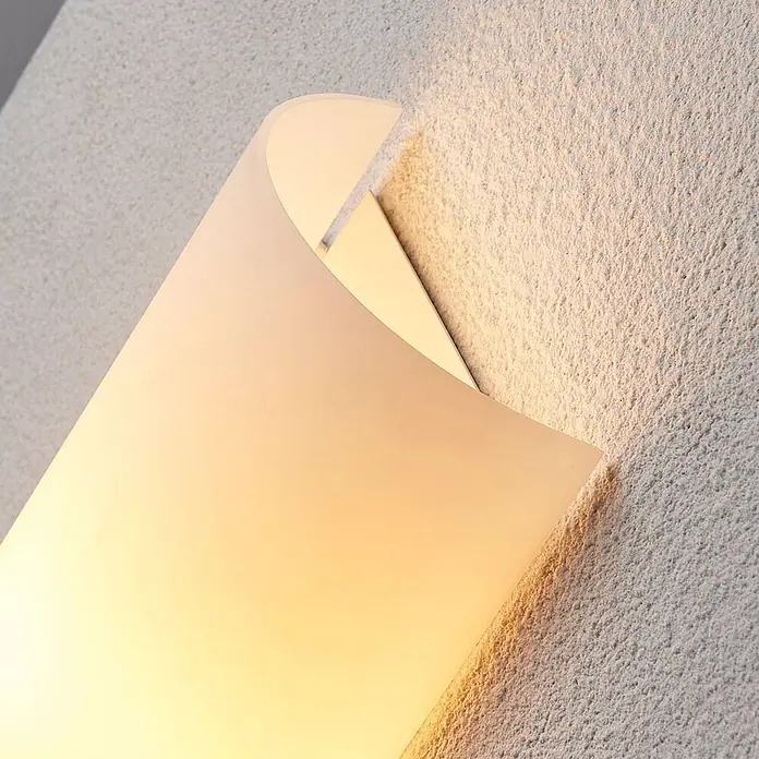 Lindby Wandleuchte Tuli - L x B x H: 7,4 x 15 x 28 cm, weiß / opal, E14 - Gebogene, mattweiße Wandleuchte, die eine strukturierte Wand mit warmem Licht beleuchtet.