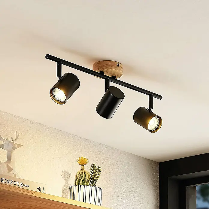 Lindby Deckenleuchte Xiomara - schwarz, GU10 - Schwarze Deckenstrahler-Schiene mit drei verstellbaren Spots und Holzsockel, beleuchtet ein Regal.