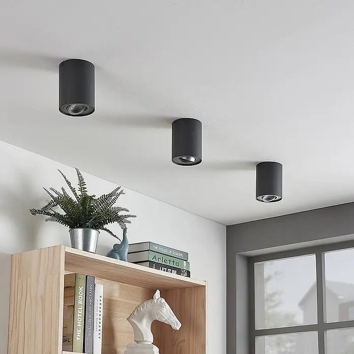 Arcchio Deckenleuchte Jolina - schwarz, GU10 - Drei schwarze zylindrische Deckenstrahler sind an einer weißen Decke montiert und beleuchten einen Raum mit Holzregal.