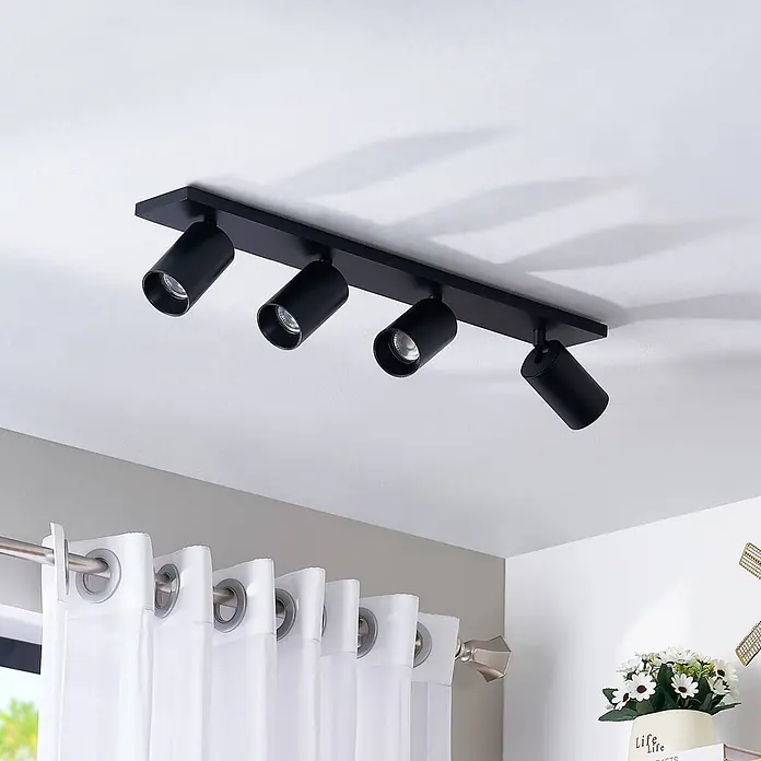 Arcchio Deckenstrahler Brinja - schwarz, schwarz, GU10 - Eine schwarze Deckenstrahlerleiste mit vier verstellbaren Spots ist an einer weißen Decke montiert und beleuchtet einen Raum mit weißen Vorhängen.
