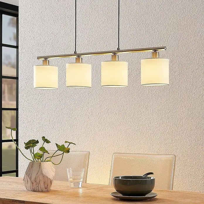 Lindby Pendelleuchte Stannis - L x B x H: 75 x 12 x 13,5 cm, weiß / opal, nickel matt, weiß, E14 - Gebürstete messingfarbene vierflammige Pendelleuchte mit Stoffschirmen über einem Esstisch aus Holz.