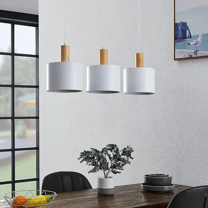 Lindby Pendelleuchte Kadir - L x B x H: 73 x 20 x 15 cm, weiß, holz, E27 - Drei moderne Pendelleuchten mit weißen Metallschirmen und Holzdetails hängen über einem Esstisch.