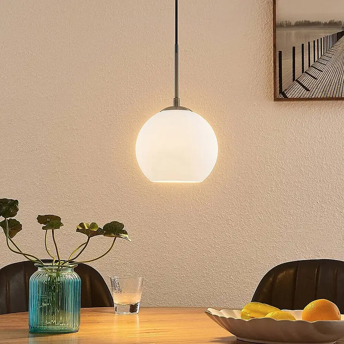 Lindby Pendelleuchte Jurian - L x B x H: 20 x 20 x 17,5 cm, weiß / opal, nickel, opal matt, E27 - Eine kugelförmige Pendelleuchte aus weißem Glas hängt über einem Holztisch und spendet Umgebungslicht.