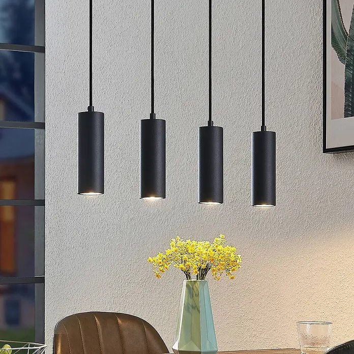 Lindby Pendelleuchte Maniva - L x B x H: 60 x 8 x 18 cm, schwarz, holz hell, Schwarz, GU10 - Vier schwarze zylindrische Pendelleuchten hängen über einem Esstisch und strahlen warmes Licht nach unten ab.
