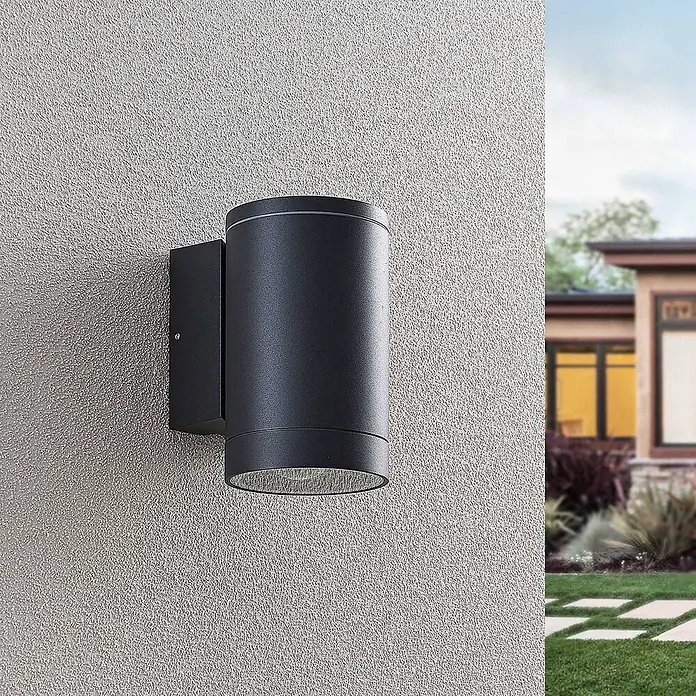 Lucande Außenwandleuchte Thomke - alu / grau / zink, alu / grau / zink, IP65 - Zylindrische schwarze Außenwandleuchte an einer strukturierten Wand, mit moderner Hausfassade und Gartenweg im Hintergrund.