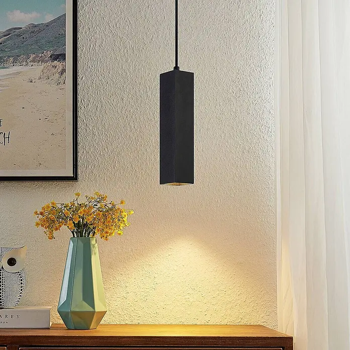 Prios Pendelleuchte Neliyah - L x B x H: 5,6 x 5,6 x 25 cm, schwarz, sandSchwarz, GU10 - Eine schwarze rechteckige Pendelleuchte beleuchtet eine strukturierte Wand über einer Holzoberfläche mit gelben Blumen in einer Vase.