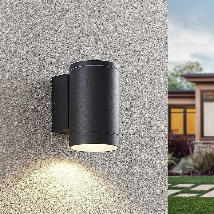 Lucande Außenwandleuchte Thomke - alu / grau / zink, alu / grau / zink, IP65 - Schwarze zylindrische Außenwandleuchte an texturierter Wand, die warmes Licht nach unten wirft, mit unscharfem Haus im Hintergrund.