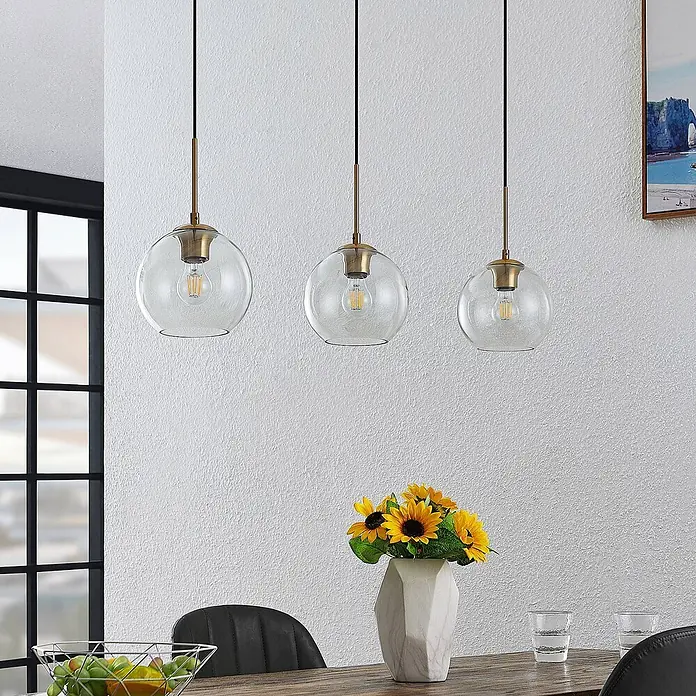 Lindby Pendelleuchte Grady - L x B x H: 110 x 20 x 17,5 cm, klar / transparent, messing, transparent, Schwarz, E27 - Drei Pendelleuchten mit klaren Glaskugeln und messingfarbenen Details, die über einem Esstisch mit Blumen hängen.