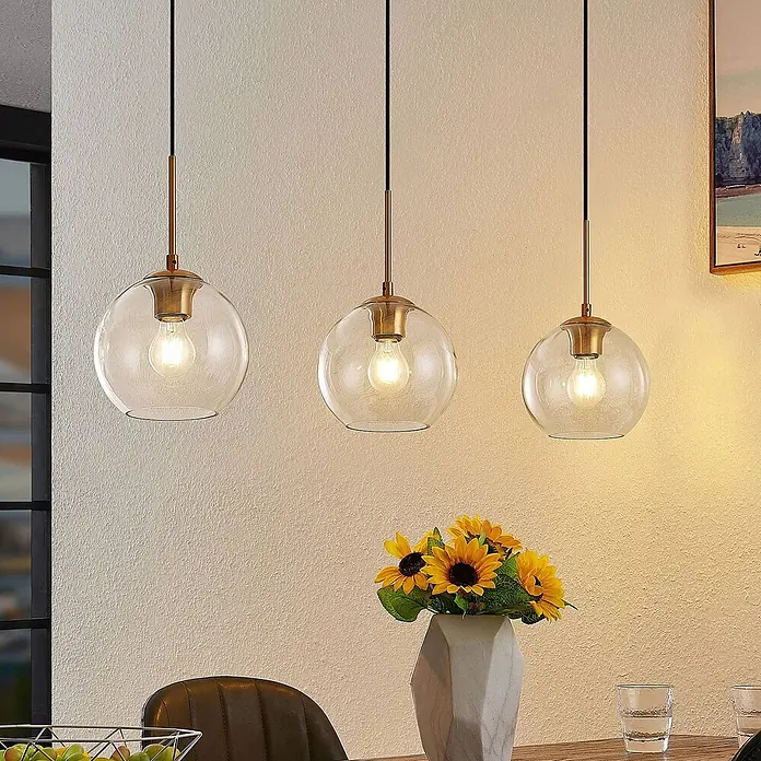 Lindby Pendelleuchte Grady - L x B x H: 110 x 20 x 17,5 cm, klar / transparent, messing, transparent, Schwarz, E27 - Drei klare Glaskugel-Pendelleuchten mit Messingakzenten, leuchtend über einem Esstisch mit Sonnenblumen.