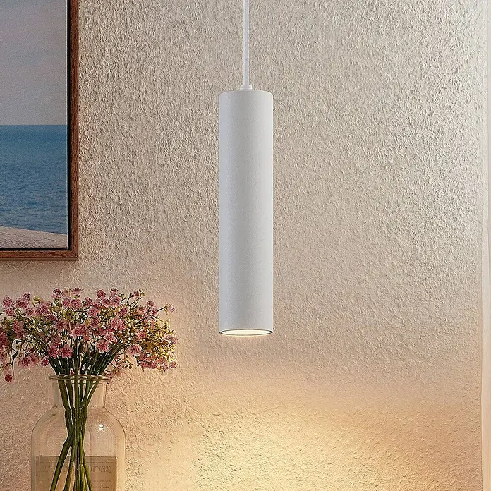 Prios Pendelleuchte Neliyah - weiß / opal, sandweiß, GU10 - Weiße zylindrische Pendelleuchte hängt und wirft einen warmen Schein auf eine strukturierte Wand und Blumendekoration.