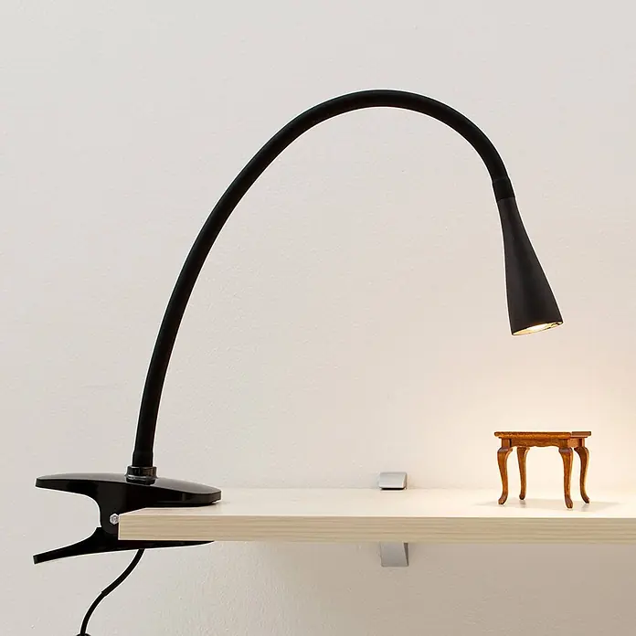 Lindby Klemmleuchte Baris - Farbe Gehäuse: schwarz - Schwarze Klemm-Schreibtischlampe mit Schwanenhalsarm, die einen Miniaturtisch auf einem hellen Holzregal beleuchtet.