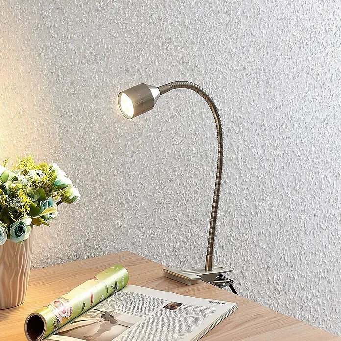 Lindby Klemmleuchte Djamila - L x B x H: 23 x 4 x 39 cm, Farbe Gehäuse: alu / grau / zink - Klemmleuchte aus gebürstetem Metall mit flexiblem Schwanenhals, beleuchtet ein Magazin auf einem Holztisch.