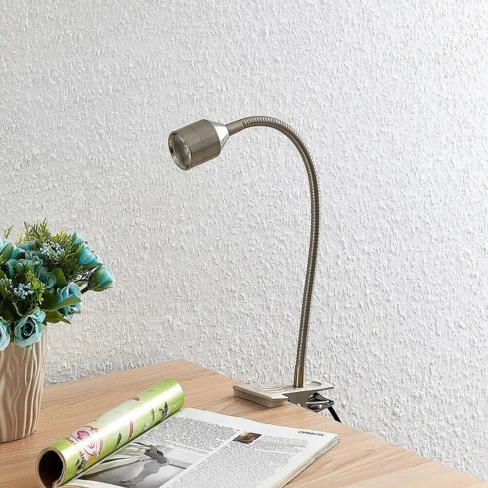 Lindby Klemmleuchte Djamila - L x B x H: 23 x 4 x 39 cm, Farbe Gehäuse: alu / grau / zink - Klemmbare Schreibtischlampe aus gebürstetem Metall mit flexiblem Schwanenhals, an einem Holztisch neben Magazin und Blumen geklemmt.