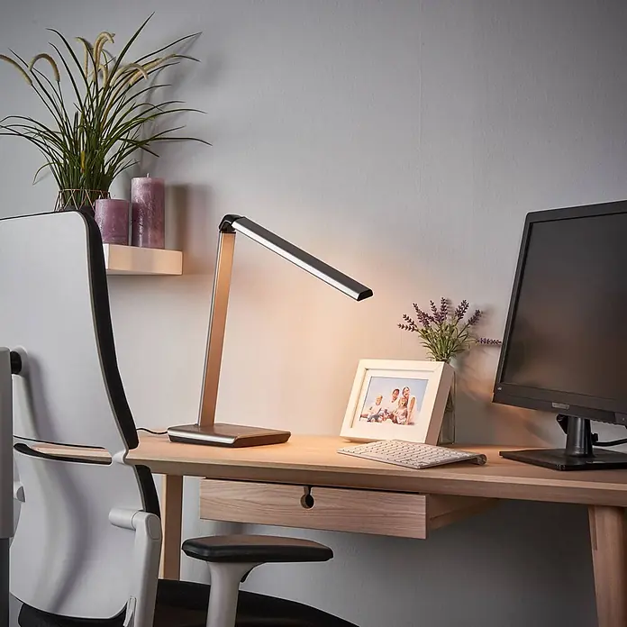 Lindby Tischleuchte Kuno - L x B x H: 37,6 x 15,8 x 46 cm, alu / grau / zink, alu / grau / zink - Eine silberne LED-Schreibtischlampe beleuchtet einen Holzschreibtisch mit Computermonitor, Tastatur und Bilderrahmen.