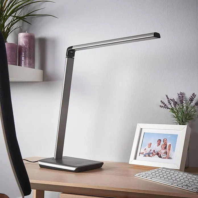 Lindby Tischleuchte Kuno - L x B x H: 37,6 x 15,8 x 46 cm, alu / grau / zink, alu / grau / zink - Moderne silbergraue LED-Schreibtischlampe mit verstellbarem Arm, auf einem Holzschreibtisch neben Tastatur und Bilderrahmen.
