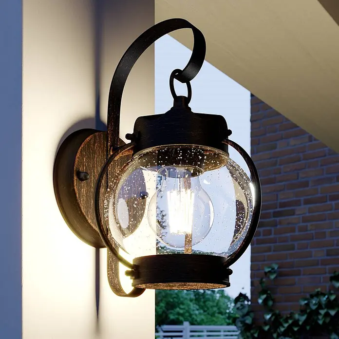 Lindby Außenwandleuchte Gerlo - braun / rost, IP23 - Dunkle Bronze-Außenwandlaterne mit blasenbesetztem Glasglobus und sichtbarer Edison-Glühbirne, an der Gebäudeaußenwand montiert.