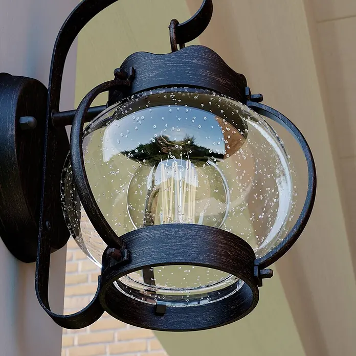 Lindby Außenwandleuchte Gerlo - braun / rost, IP23 - Rostbraune Außenwandleuchte mit strukturiertem Glas und sichtbarer Edison-LED-Lampe, an einem Gebäude montiert.
