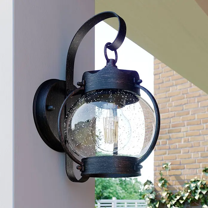 Lindby Außenwandleuchte Gerlo - braun / rost, IP23 - Dunkelbronze Außenwandleuchte mit klarem, blasigem Glas und sichtbarer Glühbirne, an cremefarbener Wand montiert.