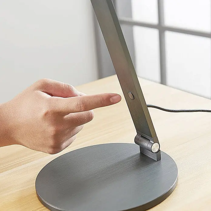 Lucande Tischleuchte Mion - L x B x H: 35 x 18 x 41 cm, alu / grau / zink, alu / grau / zink - Dunkelgraue Schreibtischlampe aus gebürstetem Metall auf einem Holztisch, eine Hand drückt ihren Bedienknopf.