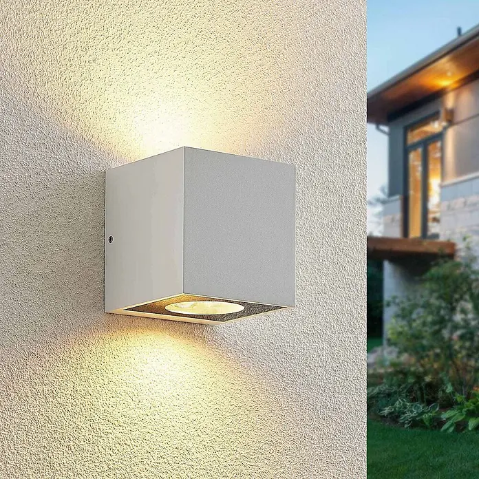 Arcchio Außenwandleuchte Tassnim - weiß / opal, weiß / opal, IP65 - Graue, würfelförmige Außenwandleuchte an einer strukturierten Wand, die warmes Licht nach oben und unten abstrahlt und das Gebäude beleuchtet.
