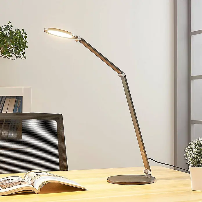 Lucande Tischleuchte Mion - L x B x H: 35 x 18 x 41 cm, alu / grau / zink, alu / grau / zink - Verstellbare LED-Schreibtischlampe in metallischem Dunkelgrau, mit ovalem, beleuchtetem Kopf, auf einem Holzschreibtisch.