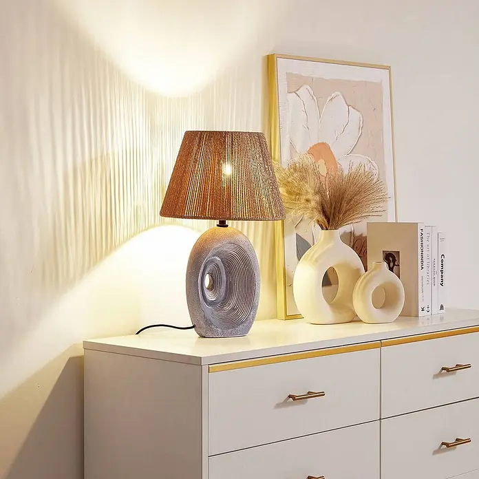 Lindby Tischleuchte Seasur - L x B x H: 29 x 29 x 48 cm, alu / grau / zink, braun / rost, E27 - Eine Tischlampe mit braunem geflochtenem Schirm und strukturiertem hellgrauem Keramiksockel, die eine Kommode beleuchtet.