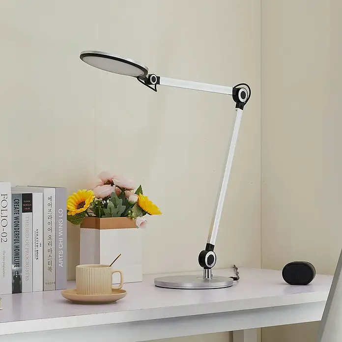 Lindby Tischleuchte Nyxaris - L x B x H: 55,7 x 20,1 x 49,7 cm, alu / grau / zink, alu / grau / zink - Eine silberne und schwarze verstellbare LED-Schreibtischlampe beleuchtet einen weißen Schreibtisch mit Büchern, Blumen, einer Tasse und einem kleinen Lautsprecher.