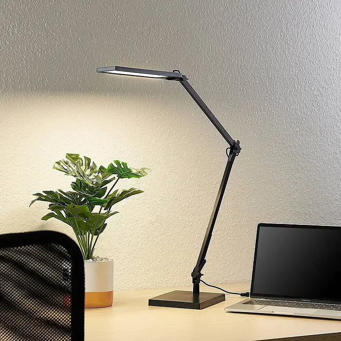 Lindby Tischleuchte Antisa - L x B x H: 65 x 14 x 47 cm, schwarz, schwarz - Schwarze, verstellbare LED-Schreibtischlampe beleuchtet einen Schreibtisch mit Pflanze und Laptop.