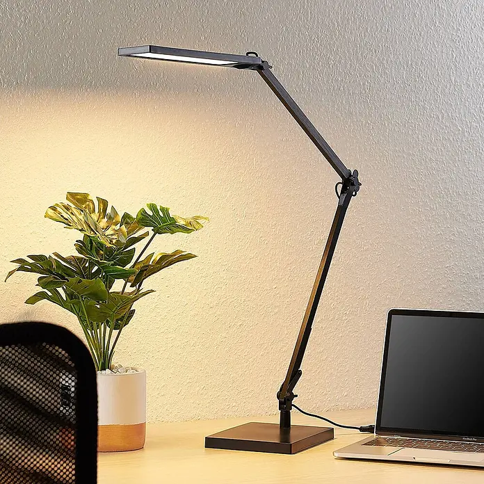 Lindby Tischleuchte Antisa - L x B x H: 65 x 14 x 47 cm, schwarz, schwarz - Schwarze, verstellbare LED-Schreibtischlampe beleuchtet einen Holzschreibtisch mit Pflanze und Laptop.