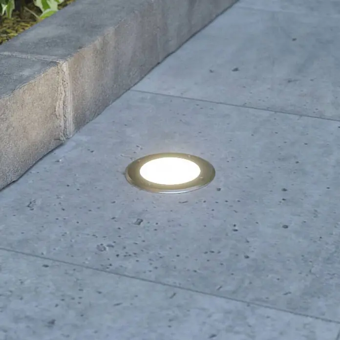 Lucande LED-Gartenstrahler Doris - 3.000 K, 12 x 12 x 15 cm - Runde, silberne Bodenleuchte mit hellem weißem Licht, in Betonplatten neben einer Steinbegrenzung eingelassen.
