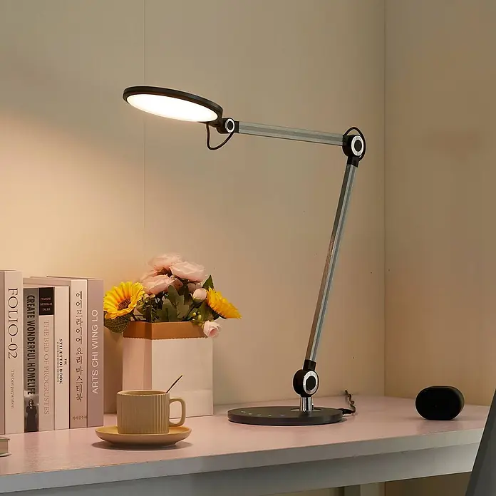 Lindby Tischleuchte Nyxaris - alu / grau / zink, alu / grau / zink - Moderne schwarz-silberne LED-Schreibtischlampe mit ovalem Kopf und Gelenkarm, die einen Schreibtisch mit Büchern und Blumen beleuchtet.