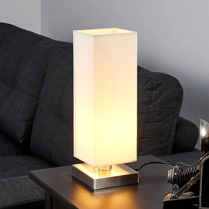 Lindby Tischleuchte Martje - L x B x H: 12 x 12 x 34,5 cm, creme / amber, E14 - Rechteckige Tischlampe mit weißem Stoffschirm und gebürstetem Metallsockel, auf einem dunklen Holztisch.