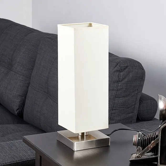 Lindby Tischleuchte Martje - L x B x H: 12 x 12 x 34,5 cm, creme / amber, E14 - Rechteckige Tischlampe mit weißem Stoffschirm und gebürstetem Metallsockel auf dunklem Beistelltisch neben grauem Sofa.