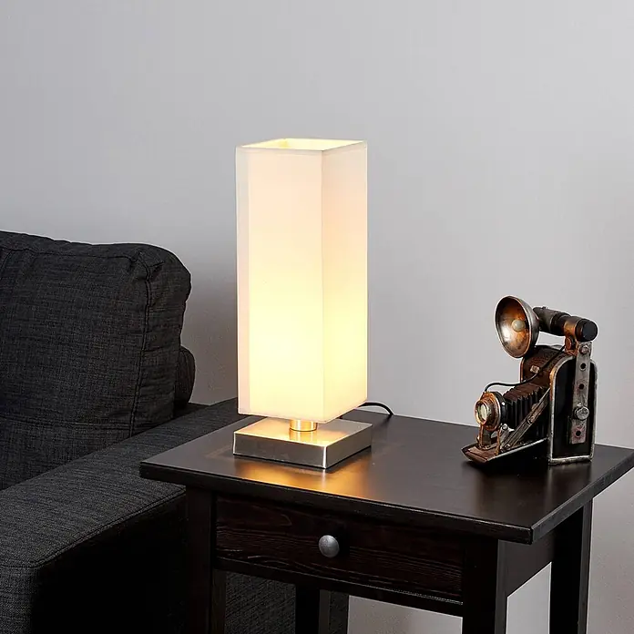 Lindby Tischleuchte Martje - L x B x H: 12 x 12 x 34,5 cm, creme / amber, E14 - Rechteckige Tischlampe mit weißem Stoffschirm und gebürsteter Metallbasis auf einem dunklen Beistelltisch neben einem grauen Sofa und einer Vintage-Kamera.