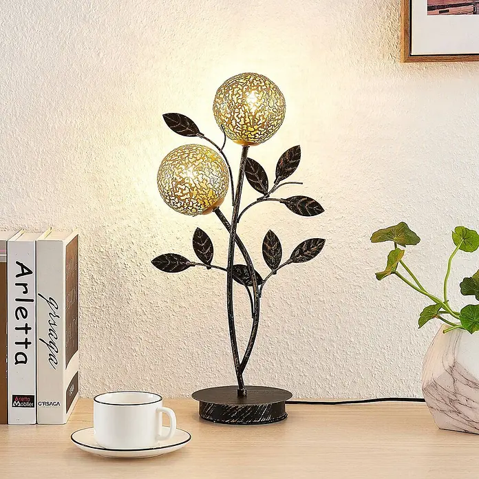 Lucande Tischleuchte Evory - schwarz, messing / gold, G9 - Dekorative Tischleuchte in dunklem Metallbaumzweigdesign mit zwei beleuchteten goldenen Filigrankugeln und Blättern auf einem Holztisch.