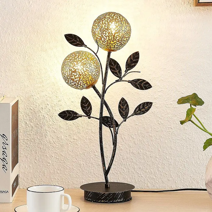 Lucande Tischleuchte Evory - schwarz, messing / gold, G9 - Dekorative Metall-Tischlampe mit zwei leuchtenden, perforierten Goldkugeln und dunklen Blättern auf einem Holztisch.