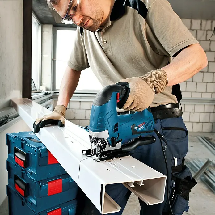 Bosch Professional Elektro-Stichsäge GST 90 BE - 500 /min - 3.100 /min, 90 mm - Ein Handwerker schneidet ein weißes Metallrohr mit einer blauen Bosch Stichsäge. Schutzbrille und Arbeitshandschuhe sind getragen.