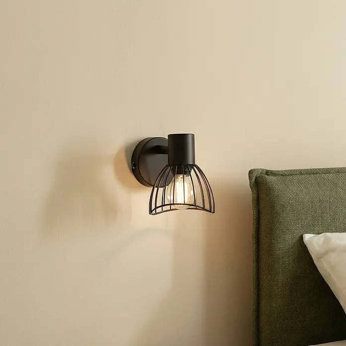 Lindby Deckenleuchte Piena - schwarz, schwarz, E14 - Schwarze Wandleuchte aus Metall mit Drahtkäfigschirm und freiliegender Glühbirne, an einer Wand neben einem Bett montiert.