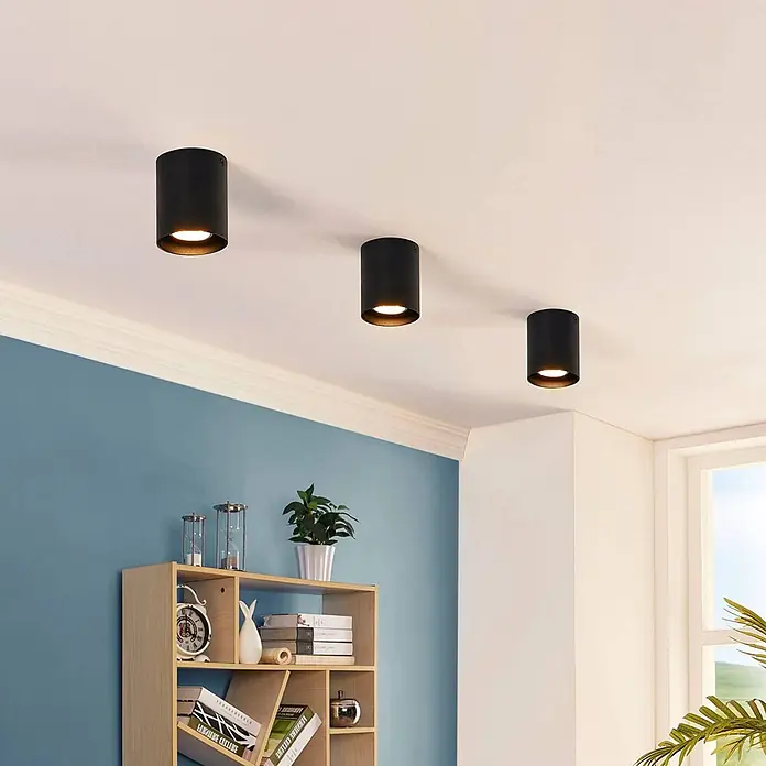 Arcchio Deckenleuchte Carson - schwarz, schwarz, GU10 - Drei schwarze zylindrische Deckenstrahler an einer weißen Decke beleuchten einen Raum mit blauen Wänden und Bücherregal.